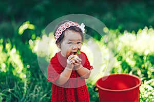 Baby girl in red dress eating pear in summer garden near red bucket