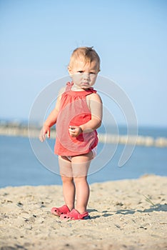 Baby girl in red dress playing on sandy beach near the sea.