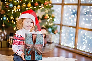 Baby girl in red santa hat holding big gift box near the Christmas tree