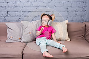 Baby girl sitting on sofa with pillows and listening to music in big headphones put on head.