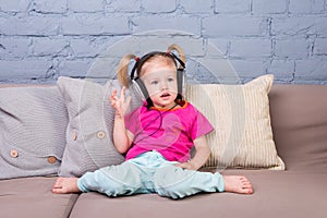 Baby girl sitting on sofa with pillows and listening to music in big headphones put on head.