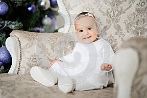 Baby girl is sitting on a vintage armchair in a white room beautiful decorated for christmas holiday with a Christmas tree and