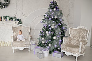 Baby girl is sitting on a vintage armchair in a white room beautiful decorated for christmas holiday with a Christmas tree and