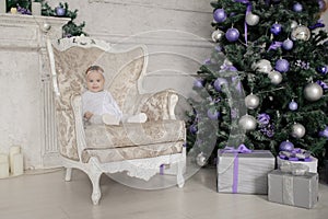 Baby girl is sitting on a vintage armchair in a white room beautiful decorated for christmas holiday with a Christmas tree and