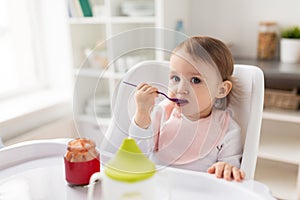 Baby girl with spoon eating puree from jar at home