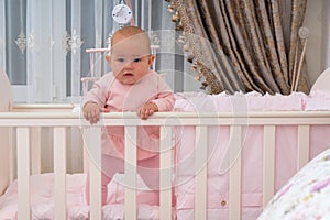 A baby girl standing in crib in pink bedroom scene.