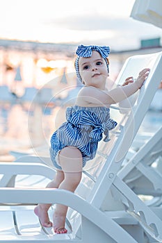 Baby girl on summer vacation, standing on lounger next to swimming pool. Sunset in the background