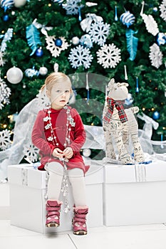 baby girl toddler with blue eyes in red dress sitting by New Year tree near deer toy