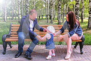 Baby girl trying to make first steps from mom to dad sitting on bench in park.