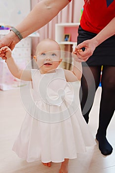 A baby girl in a white dress takes first steps, holding on to grandmother& x27;s arms