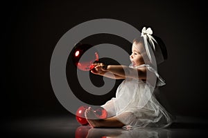 Baby girl in white headband and dress, barefoot. Holding two red balls, looking up, sitting on floor. Twilight, black background.