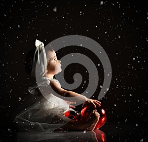 Baby girl in white headband and dress, barefoot. Holding two red balls, looking up, sitting on floor. Twilight, black background
