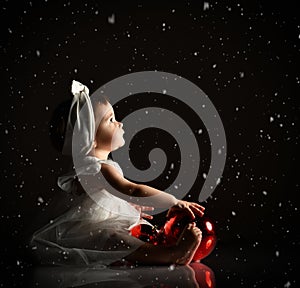 Baby girl in white headband and dress, barefoot. Holding two red balls, looking up, sitting on floor. Twilight, black background.