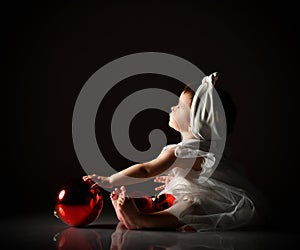 Baby girl in white headband and dress, barefoot. Holding two red balls, looking up, sitting on floor. Twilight, black background.