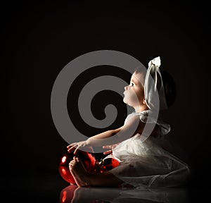 Baby girl in white headband and dress, barefoot. Holding two red balls, looking up, sitting on floor. Twilight, black background.