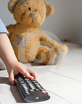 Baby hand holding the TV remote control, teddy bear in the background, selective focus