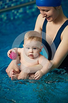 Baby holding toy balls and sitting in his mother`s hands in clear blue water of swimming pool during wellness treatments