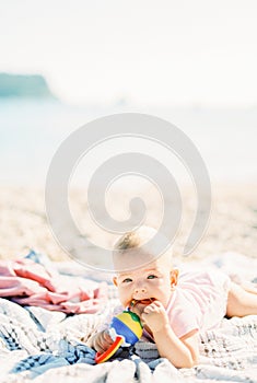 Baby lies on his tummy on the beach and gnaws on a rattle