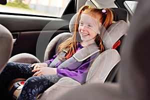 Baby little red-haired girl smiling while sitting in a child car seat.