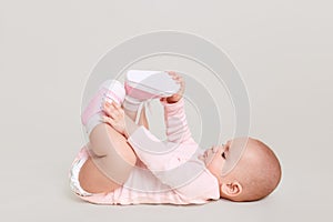 Baby lying on floor and playing with her feet, charming infant wearing pink bodysuit and shocks indoor, cute kid isolated over