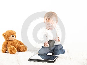 Baby with a mobile in his hand and with a tablet and a teddy bear on the floor