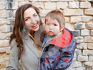 Baby with mom on a brick background. looking at the camera in warm clothes. Happy and young mother sitting on brick background and