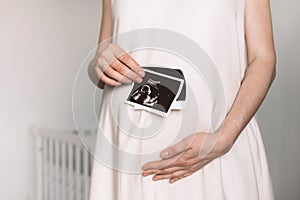 In baby nursery pregnant woman in white dress holding ultrasound image, baby sonography. Concept of pregnancy, health