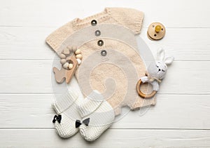 Baby pacifier, toys, knitted romper and booties on white wooden background, flat lay
