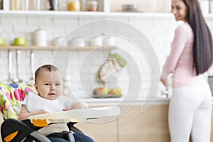 Baby playing on high chair in kitchen, mother washing dishes