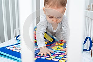 Baby playing with montessori busy book sitting in crib. Concept of smart books and modern toys