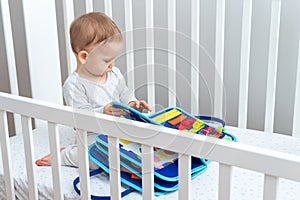 Baby playing with montessori busy book sitting in crib. Concept of smart books and modern toys