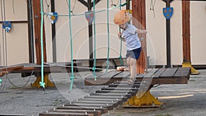 The baby is playing in a rope town on the street. A boy walks on children`s equipment and falls. A kid goes on a tottering wooden