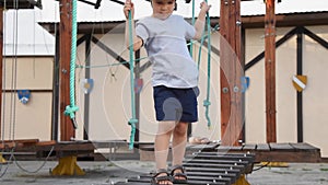 The baby is playing in a rope town on the street. A boy walks on children`s equipment. A kid goes on a tottering wooden bridge in