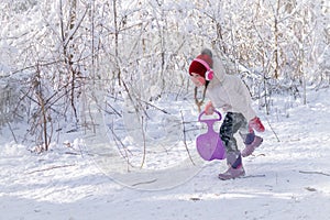 The baby is playing in a snow-covered glade. Concept of active children`s recreation. A cheerful little girl runs with an iceboat