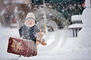 Baby playing with teddy in the snow, winter time. Little toddler boy in blue coat, holding suitcase and teddy bear, playing