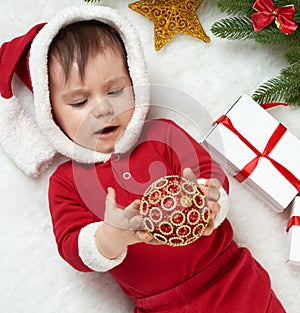 Baby portrait in christmas decoration, dressed as Santa, lie on fur near fir tree and play with gifts, winter holiday concept