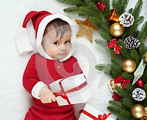 Baby portrait in christmas decoration, dressed as Santa, lie on fur near fir tree and play with gifts, winter holiday concept