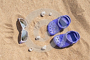 Baby purple clog and sunglasses on the sand on the beach. Cute details of summer holidays. Baby shoes on the beach.