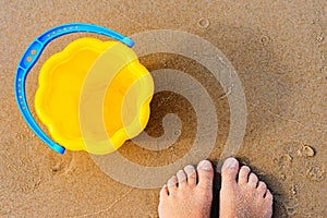 Baby`s bare feet on a golden sandy beach next to a yellow toy bucket. Vacation concept