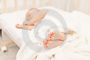 Baby`s legs in focus against the background of a sleeping baby in a white bed, a healthy and calm baby`s sleep