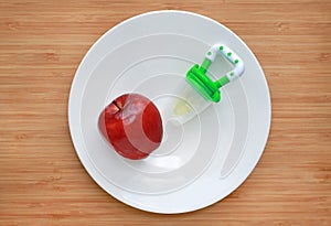 Baby`s nibbler and red apple on white plate against wooden background. Organic baby food concept photo