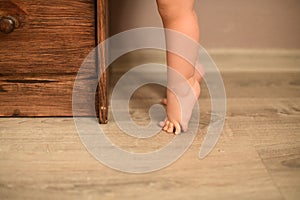 The baby& x27;s small feet are on the floor standing in tip toe in front of the dresser.