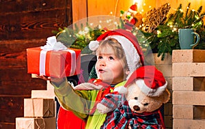 Baby Santa boy holding and looking at Christmas box gift trying to guess what is inside it. Little kid at Santa`s hat