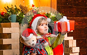 Baby Santa boy holding and looking at Christmas box gift trying to guess what is inside it. Little kid at Santa`s hat