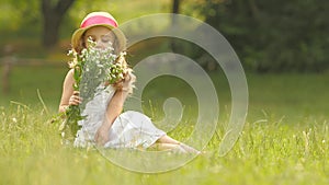 Baby sits on a glade in a sarafan and hat, holds wild flowers and smiles