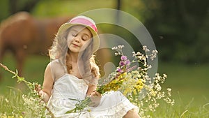 Baby sits on a glade in a sarafan and hat, holds wild flowers and smiles. Slow motion