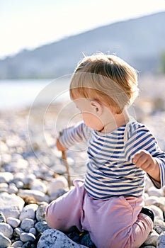 Baby sits on his knees on a pebble beach and digs the ground with a stick