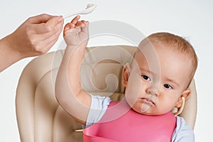 A baby sitting on a chair pushes away his mother`s hand with a spoon with food