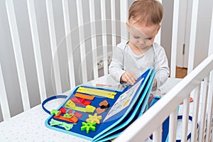 Baby sitting in a crib playing with montessori busy book. Concept of keeping children from screen by activity books and