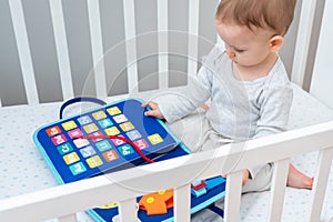 Baby sitting in a crib playing with montessori busy book. Concept of keeping children from screen by activity books and